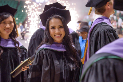 A girl wearing graduation cap and gown smiling at the camera surrounded by other people in graduation attrie with a spark behind her head